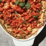 A casserole dish filled with creamy Hot Corn Dip, topped with bright red crunchy snacks and garnished with fresh cilantro leaves, sits on a sunlit surface.
