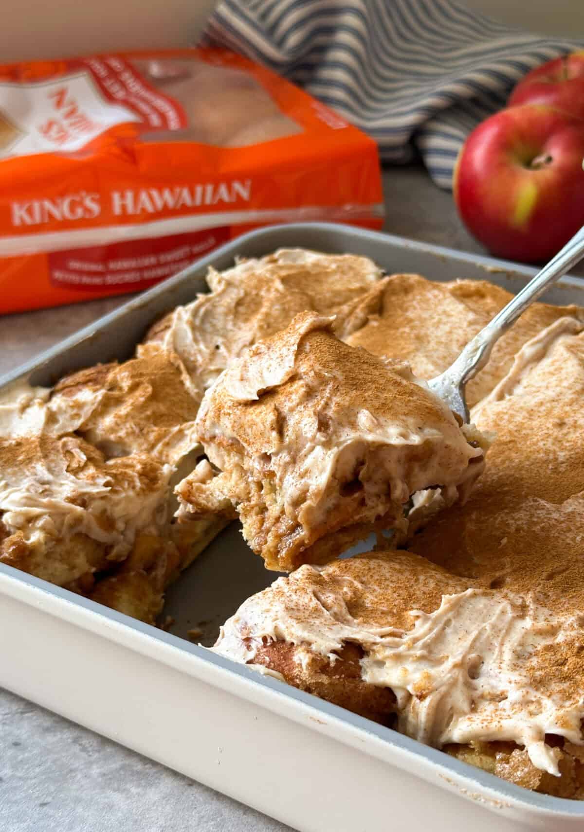 A baking dish filled with a creamy, cinnamon-topped dessert—like Apple Cinnamon Rolls—being served with a spatula. In the background, a package of King’s Hawaiian rolls and two fresh apples add to the cozy scene.