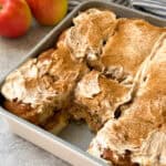 A pan of frosted Apple Cinnamon Rolls with a piece missing, topped with cinnamon, sits on a countertop near two apples and a striped cloth in the background.