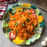 A colorful plate of tagliatelle pasta in a tomato-based sauce, garnished with fresh basil leaves, sits on a leaf-patterned dish. Nearby, a Raw Carrot Salad adds brightness alongside a red and white striped cloth, wooden utensils, and garlic bulbs.