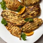 Grilled chicken breasts with visible grill marks are arranged on a white plate, garnished with fresh parsley and orange slices, featuring the zesty flavors of a Golden Citrus Chicken Marinade.