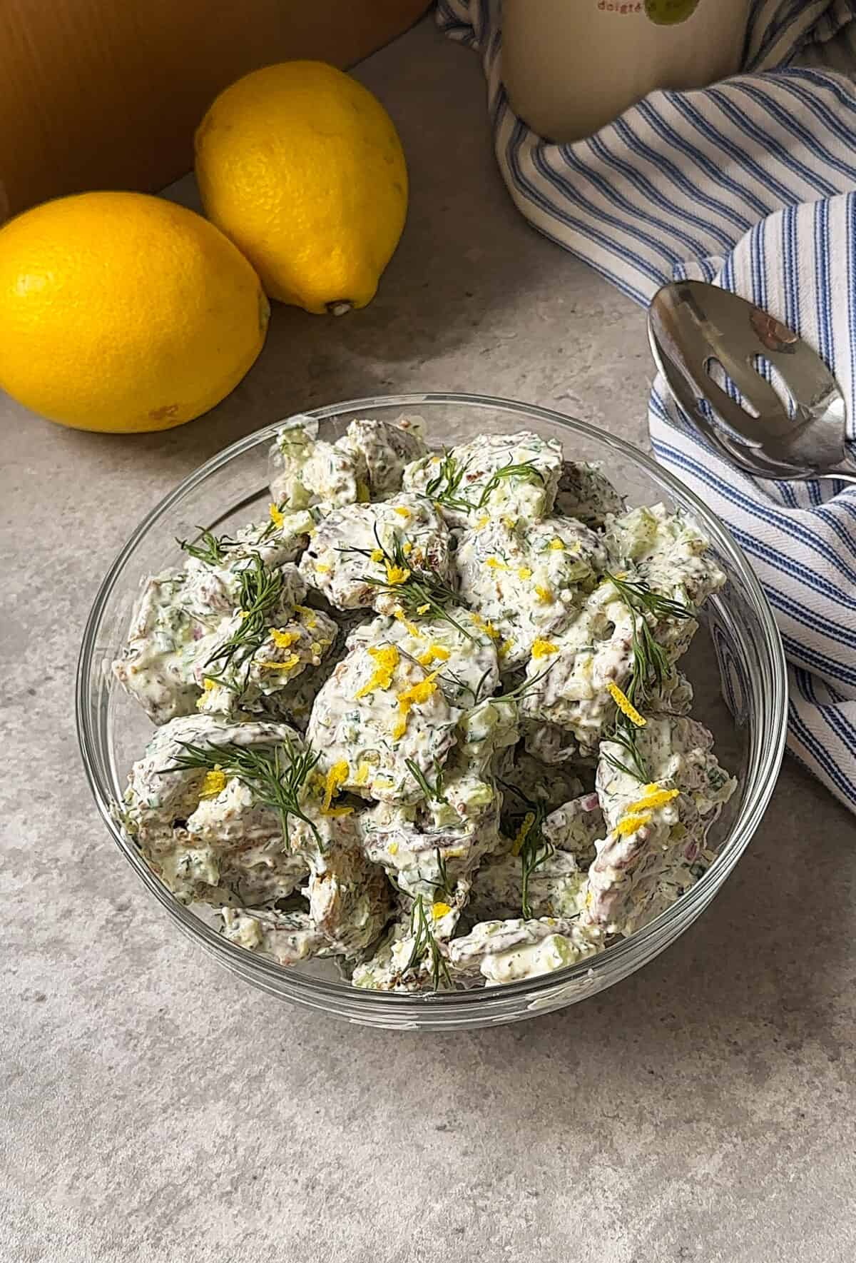 A glass bowl filled with creamy potato salad garnished with fresh dill and lemon zest sits on a gray surface near two whole lemons, a serving spoon, and a blue striped cloth—perfect alongside Crispy Smashed Potatoes for your next meal.
