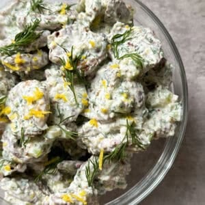 A glass bowl filled with creamy potato salad made from smashed baby potatoes, garnished with fresh dill and lemon zest, sits on a light gray surface.