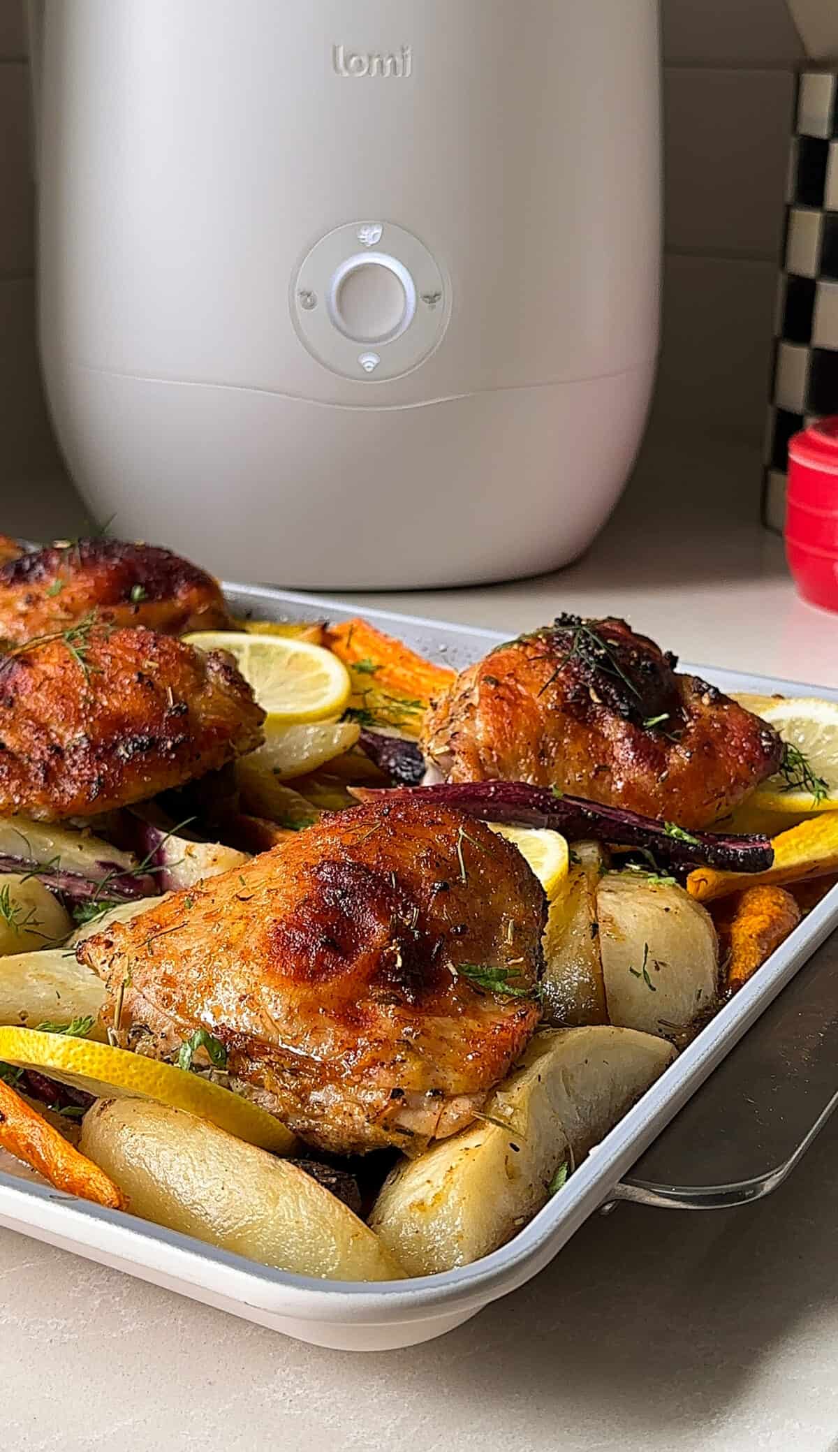 A tray of Sheet Pan Chicken and Potatoes with carrots and lemon slices sits on a kitchen counter. In the background, a white countertop appliance and checkered jar add charm to this inviting chicken and potatoes recipe scene.