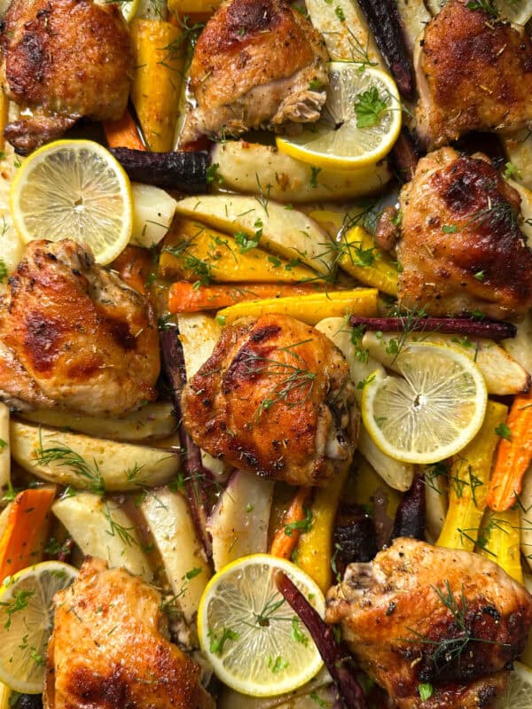 Roasted chicken thighs with golden brown skin, surrounded by colorful roasted carrots, parsnips, potatoes, fresh herbs, and lemon slices on a baking sheet—a delicious sheet pan chicken and potatoes recipe.