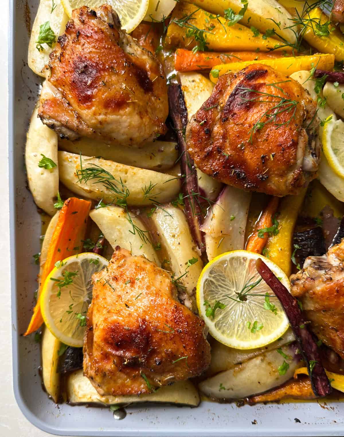 Roasted chicken thighs with golden, crispy skin are served on a baking sheet alongside colorful roasted carrots, parsnips, lemon slices, and fresh green herbs—making this a vibrant take on a classic sheet pan chicken and potatoes recipe.