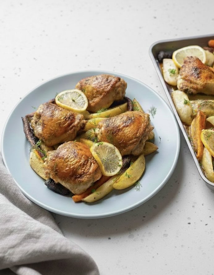 A plate of Sheet Pan Chicken and Potatoes garnished with lemon slices, served with golden potato wedges and roasted vegetables. In the background, a baking tray holds more chicken and vegetables for this delicious chicken and potatoes recipe.