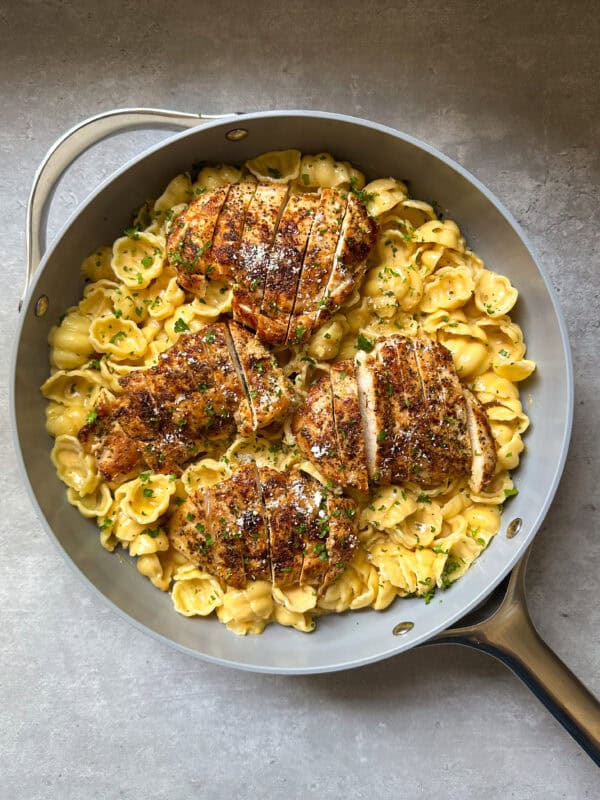 A skillet filled with creamy shell pasta topped with four pieces of seasoned, sliced chicken breast, garnished with chopped herbs and grated cheese.