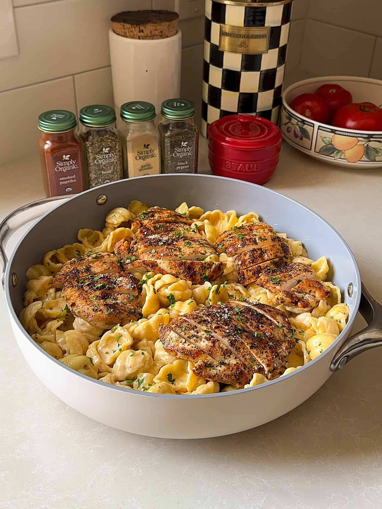 A blackened chicken Alfredo recipe skillet filled with creamy shell pasta topped with several grilled chicken breasts, sitting on a kitchen counter near spice jars, tomatoes, and a decorative canister.