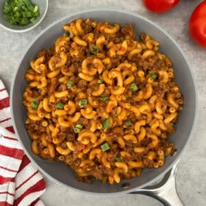 A skillet filled with macaroni pasta, ground beef, and tomato sauce creates a classic Homemade Hamburger Helper Recipe, garnished with chopped green onions. Fresh tomatoes and a bowl of green onions sit nearby with a red and white striped towel.