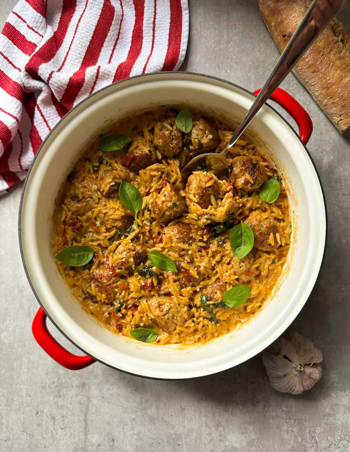 A red-handled pot filled with orzo pasta, marry me chicken meatballs, tomato sauce, and fresh basil leaves sits on a gray surface next to a striped kitchen towel, a loaf of bread, and a garlic bulb. A spoon rests in the pot.