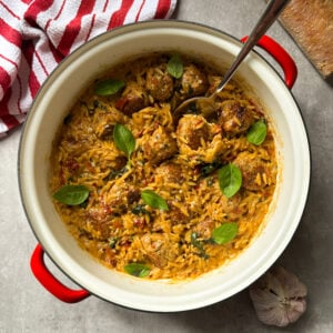 A red-handled pot filled with orzo pasta, marry me chicken meatballs, tomato sauce, and fresh basil leaves sits on a gray surface next to a striped kitchen towel, a loaf of bread, and a garlic bulb. A spoon rests in the pot.