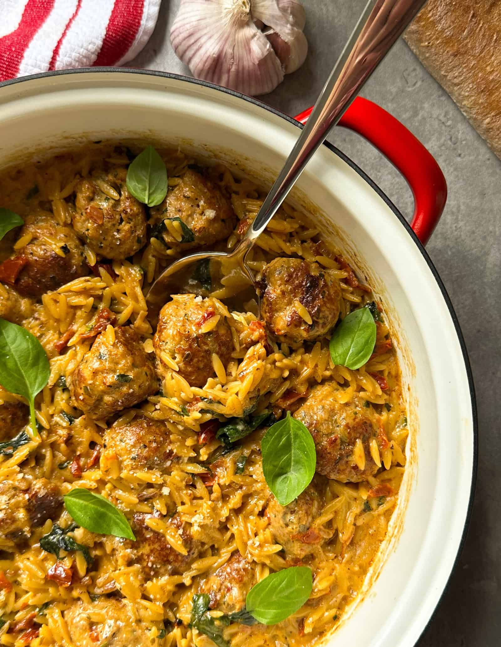 A red pot filled with creamy orzo, marry me chicken meatballs, and spinach, garnished with fresh basil leaves. A spoon rests inside the pot. Garlic and a striped towel are visible nearby on the gray countertop.