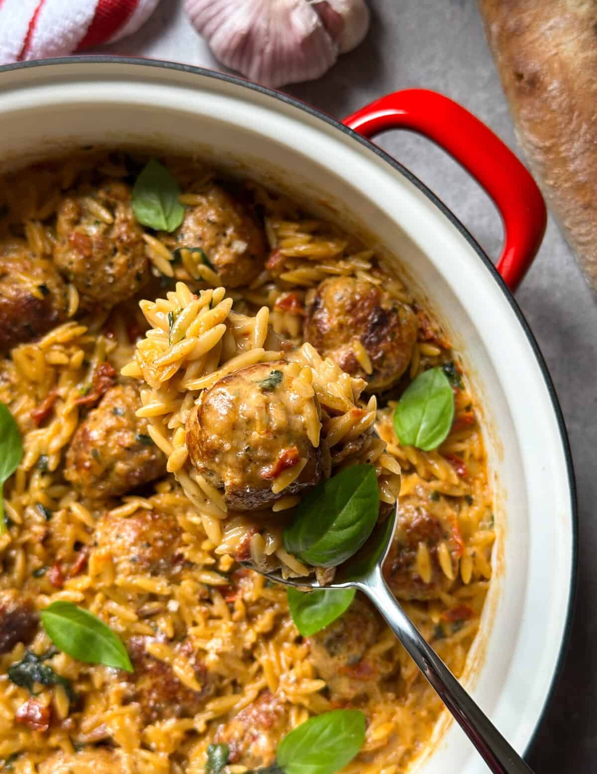 A close-up of a pot with creamy orzo pasta, marry me chicken meatballs, and fresh basil leaves. A spoon lifts a portion, showing the orzo and a meatball. Garlic cloves and bread are visible in the background.