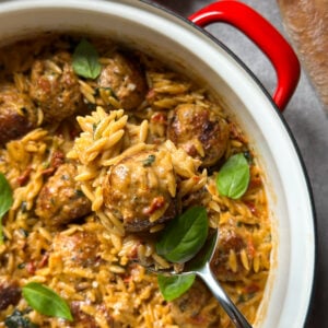A red-handled pot filled with creamy marry me chicken orzo and meatballs, garnished with fresh basil leaves. A spoon scoops up a meatball and orzo. Garlic cloves and a loaf of bread are nearby on the table.