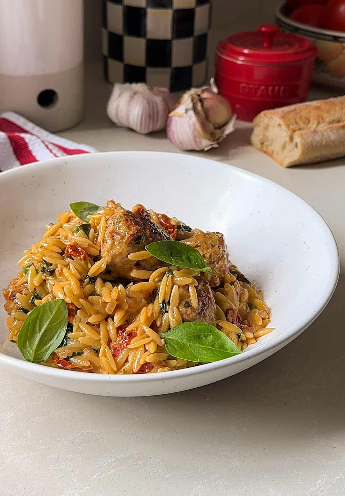 A white bowl filled with creamy orzo pasta, marry me chicken meatballs, sun-dried tomatoes, spinach, and fresh basil sits on a kitchen counter with garlic, a baguette, and kitchenware in the background.