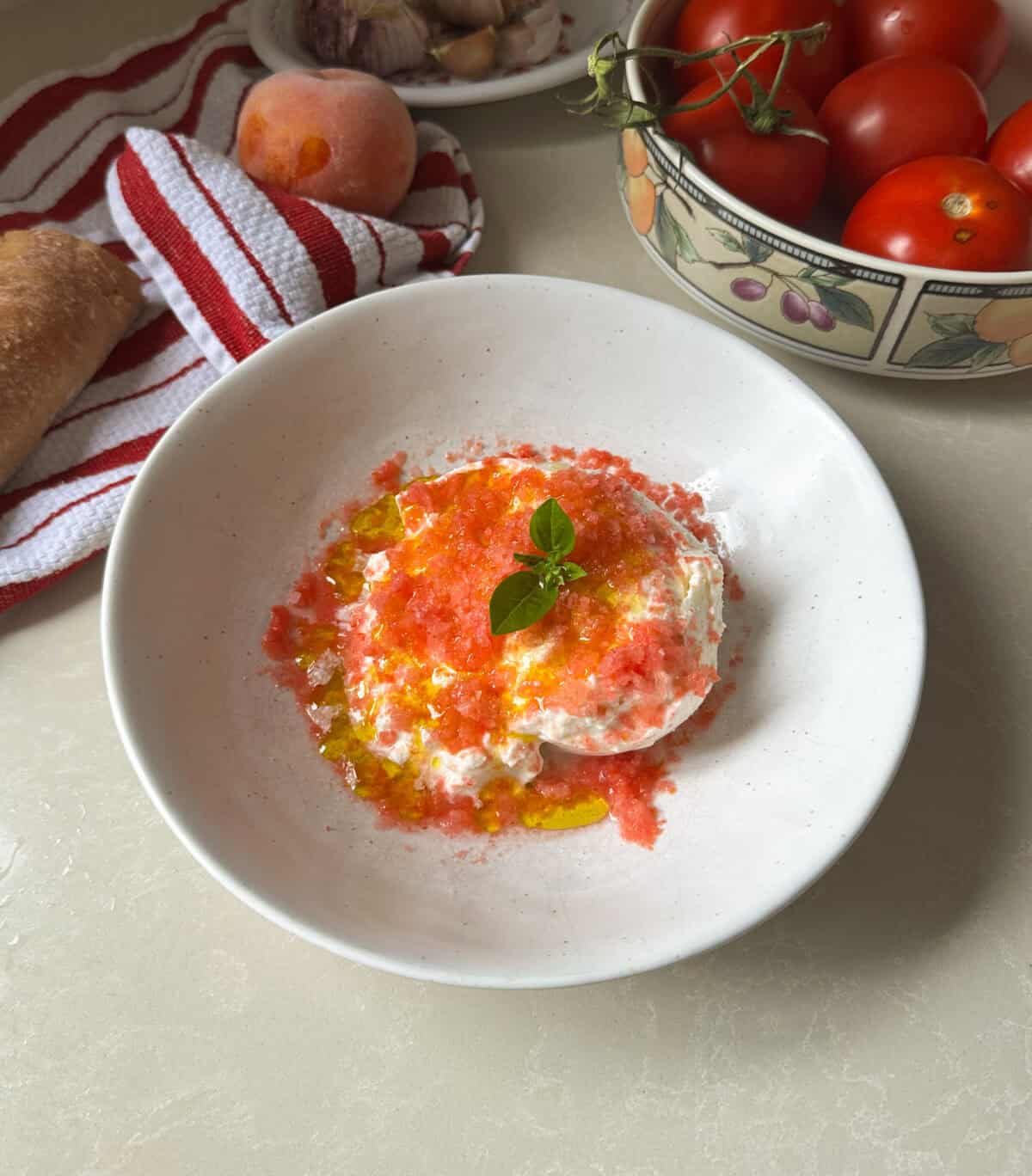 A white bowl with a burrata appetizer with shaved tomatoes, olive oil, and a basil sprig. Nearby are tomatoes on the vine, a peach, garlic, bread, and a red-striped towel.