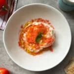 A plate of creamy burrata with shaved tomatoes, drizzled with olive oil and topped with basil, is surrounded by fresh tomatoes, bread, and a container of salt on a gray surface.