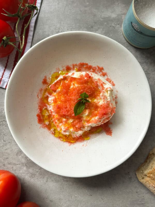 A plate of creamy burrata with shaved tomatoes, drizzled with olive oil and topped with basil, is surrounded by fresh tomatoes, bread, and a container of salt on a gray surface.