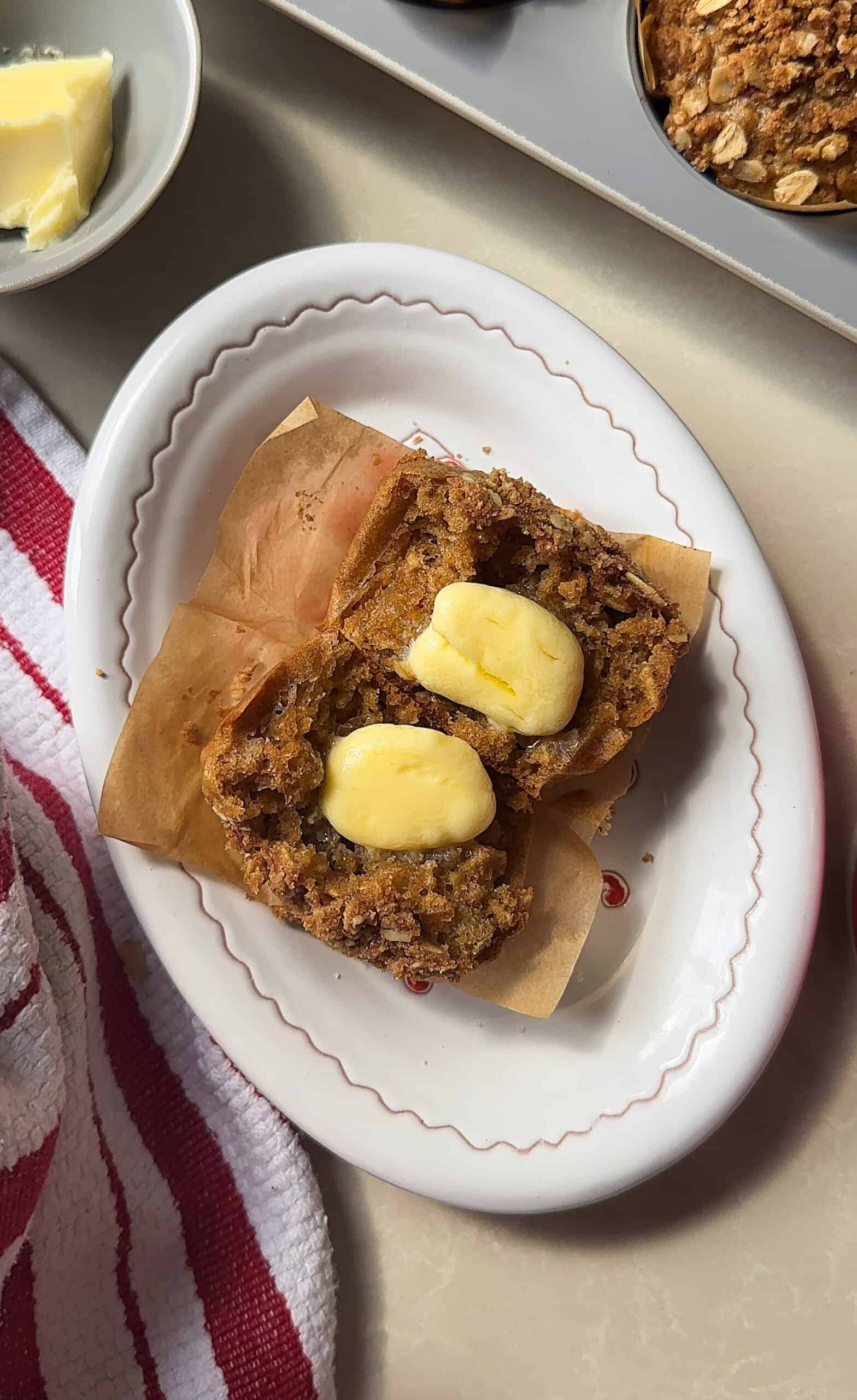 An oatmeal muffin split in half on a white plate, with pats of melting butter on each half. The muffin sits on parchment paper, and a red-striped towel is nearby. Perfect for fans of oatmeal raisin muffins.