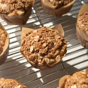 Close-up of oatmeal muffins in brown paper wrappers cooling on a metal rack, with sunlight highlighting their crumbly, golden-brown streusel topping.