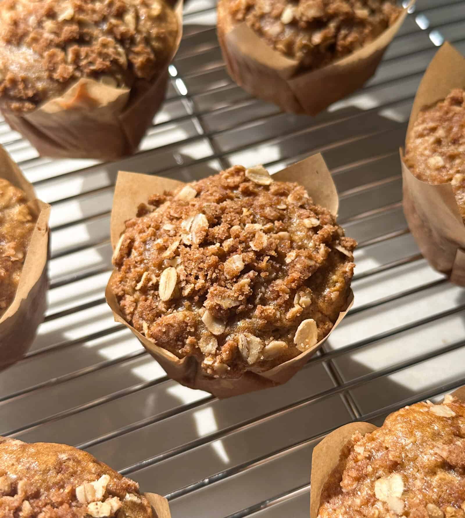 Close-up of oatmeal muffins in brown paper wrappers cooling on a metal rack, with sunlight highlighting their crumbly, golden-brown streusel topping.