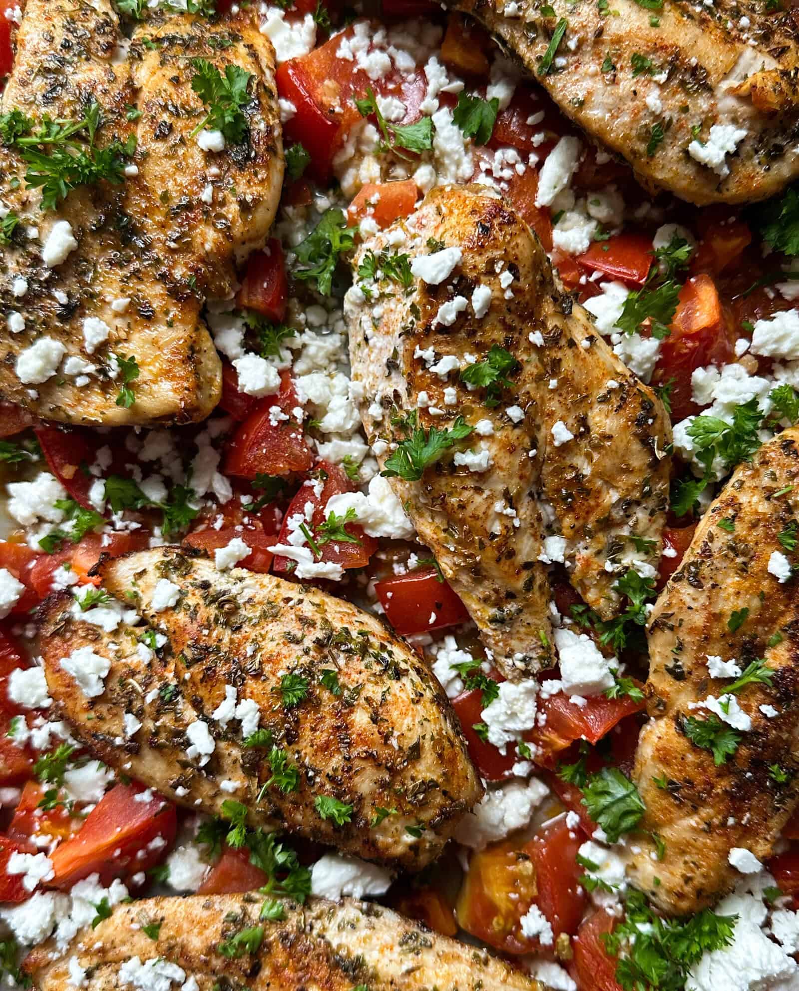 Seasoned grilled chicken breasts are served on a bed of chopped tomatoes, topped with crumbled feta and fresh parsley—a vibrant and flavorful sheet pan chicken recipe with visible herbs and spices on the chicken.
