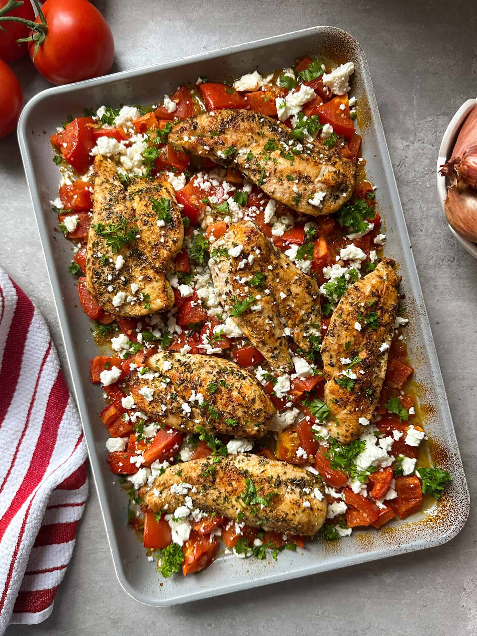 Sheet pan chicken with roasted tomatoes and feta: Seasoned baked chicken breasts, chopped tomatoes, crumbled feta, and fresh herbs. A red and white towel, tomatoes, and a shallot sit beside the pan on a gray countertop.