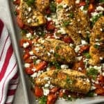 Seasoned sheet pan chicken with roasted tomatoes and feta, baked to perfection with fresh herbs. A red and white striped kitchen towel sits nearby on the countertop.