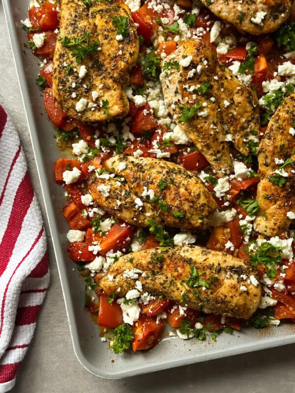 Seasoned sheet pan chicken with roasted tomatoes and feta, baked to perfection with fresh herbs. A red and white striped kitchen towel sits nearby on the countertop.