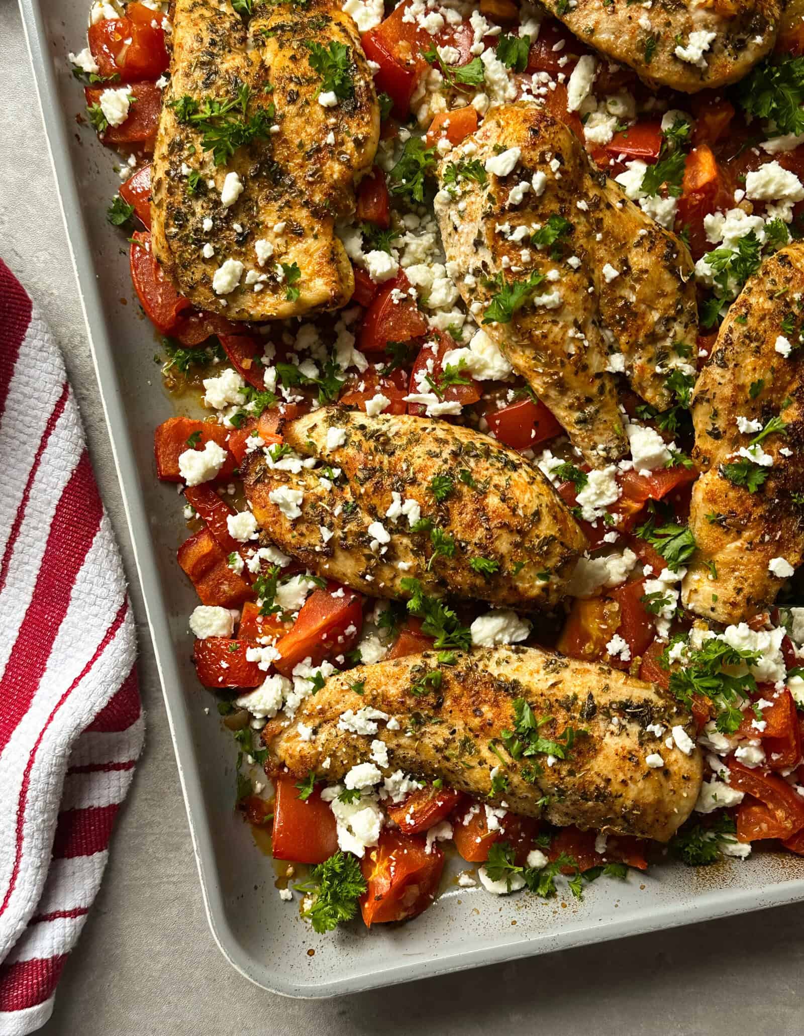 Seasoned sheet pan chicken with roasted tomatoes and feta, baked to perfection with fresh herbs. A red and white striped kitchen towel sits nearby on the countertop.