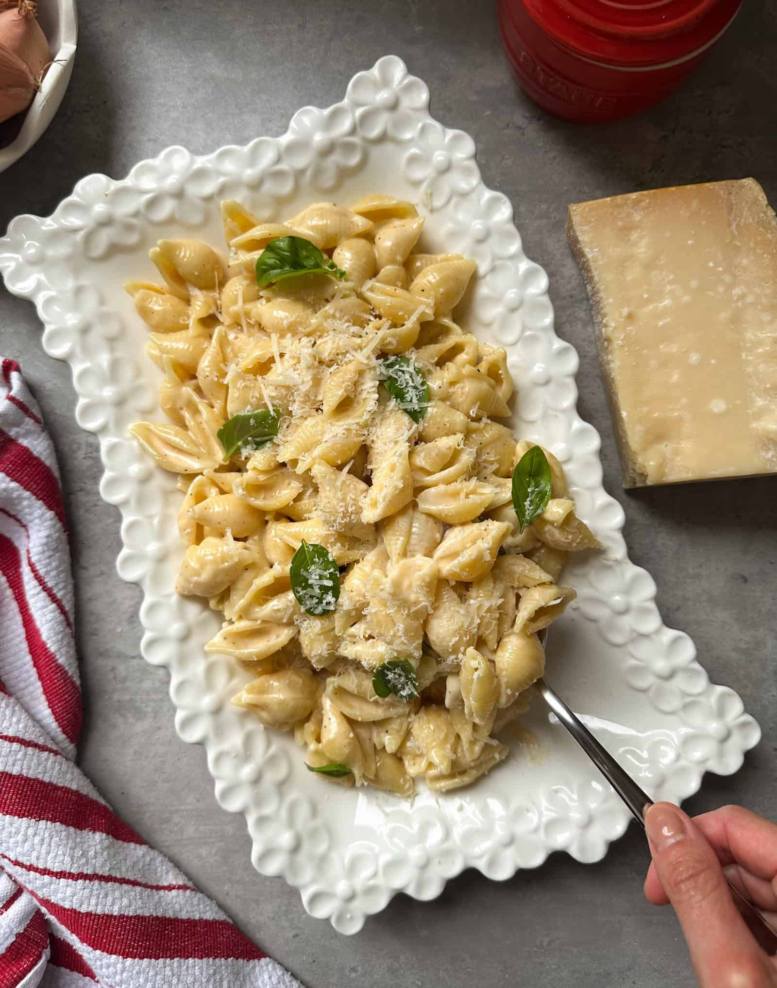A rectangular white plate with shell pasta tossed in a garlic cream sauce for pasta, topped with grated cheese and basil leaves. A hand holds a fork at the edge. Nearby are a red and white striped towel, a wedge of cheese, and a red container on a gray surface.