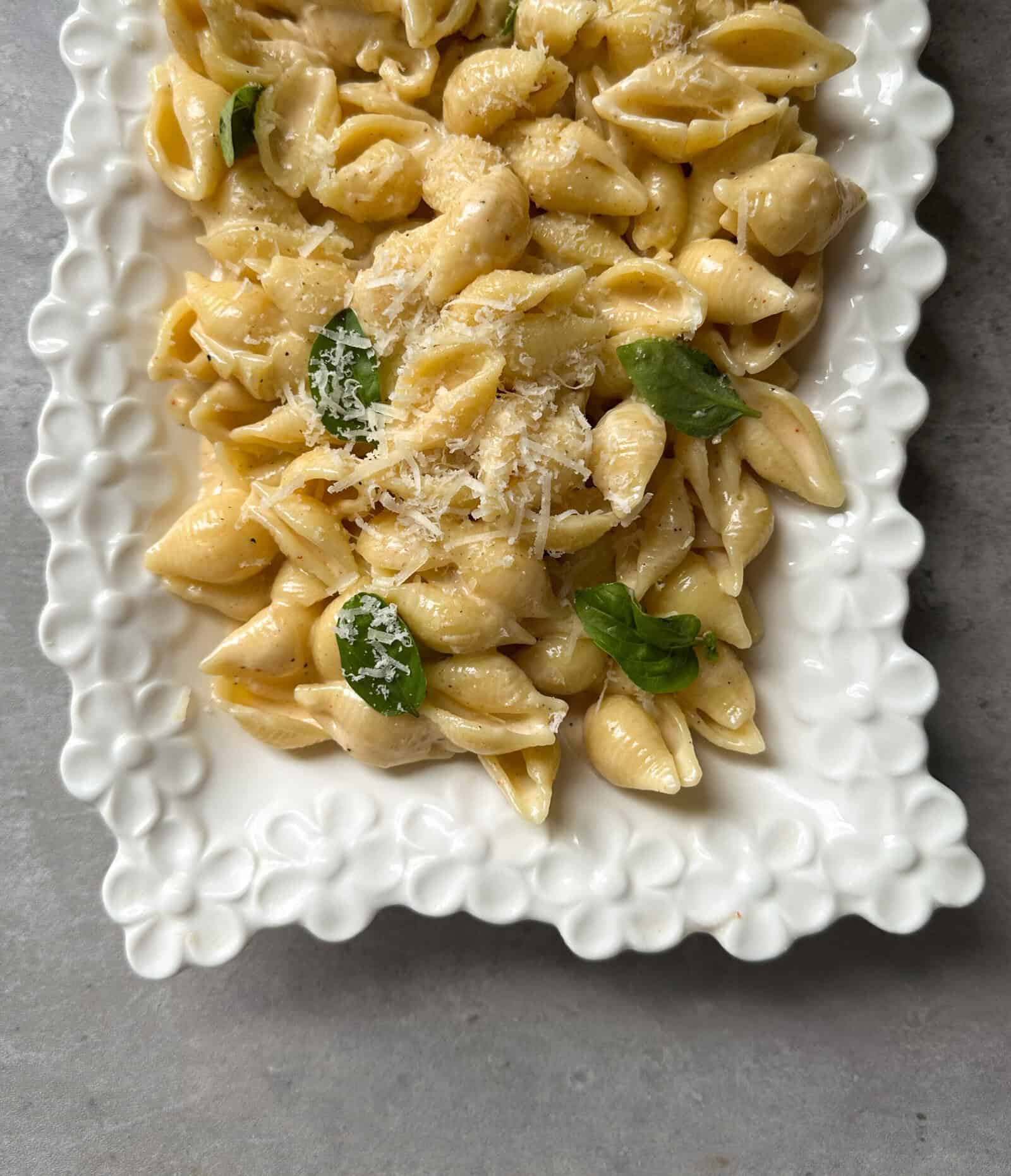 Pasta shells topped with grated cheese and fresh basil leaves, served on a decorative white plate with a scalloped edge, all complemented by a rich garlic cream sauce for pasta, set on a gray surface.