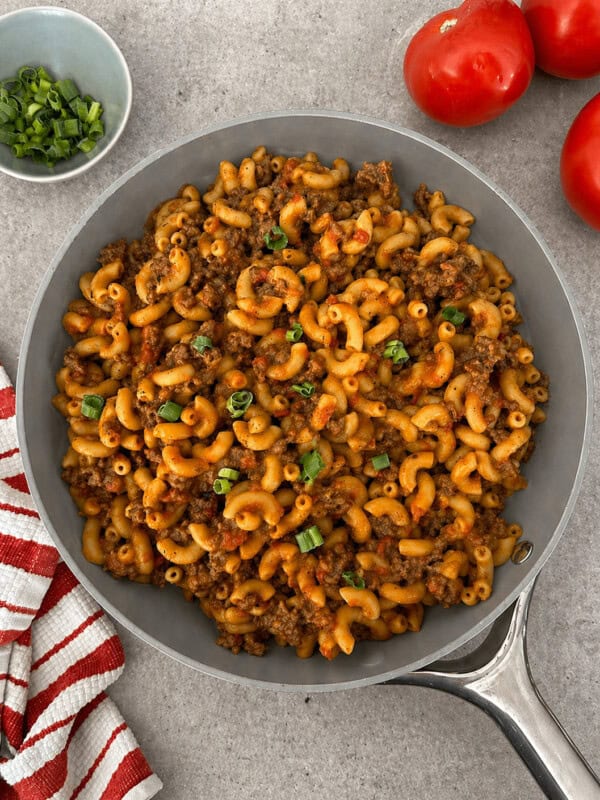 A skillet filled with macaroni, ground beef, and sauce, garnished with chopped green onions creates the ultimate Homemade Hamburger Helper Recipe. Next to it are fresh tomatoes, a bowl of green onions, and a red-striped towel on a gray surface.
