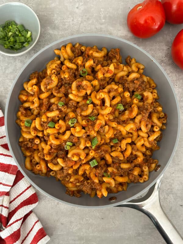 A skillet filled with macaroni pasta mixed with ground beef and tomato sauce, garnished with chopped green onions—this is a classic Homemade Hamburger Helper Recipe. Nearby are a bowl of green onions, tomatoes, and a red-and-white striped towel.