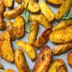 Golden, roasted potato wedges scattered on a baking sheet, garnished with fresh rosemary sprigs. These crispy roasted potatoes have a seasoned exterior and a warm, inviting appearance—perfect for anyone seeking the ultimate crispy roasted potatoes recipe.