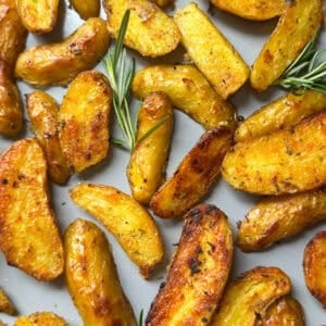 Golden, roasted potato wedges scattered on a baking sheet, garnished with fresh rosemary sprigs. These crispy roasted potatoes have a seasoned exterior and a warm, inviting appearance—perfect for anyone seeking the ultimate crispy roasted potatoes recipe.