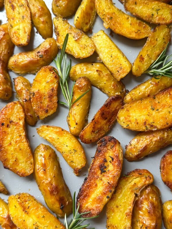 Golden, crispy roasted potatoes are scattered on a baking tray, garnished with fresh rosemary. Perfectly seasoned with a slightly charred look, they’re an irresistible example of how to make crispy roasted potatoes at home.