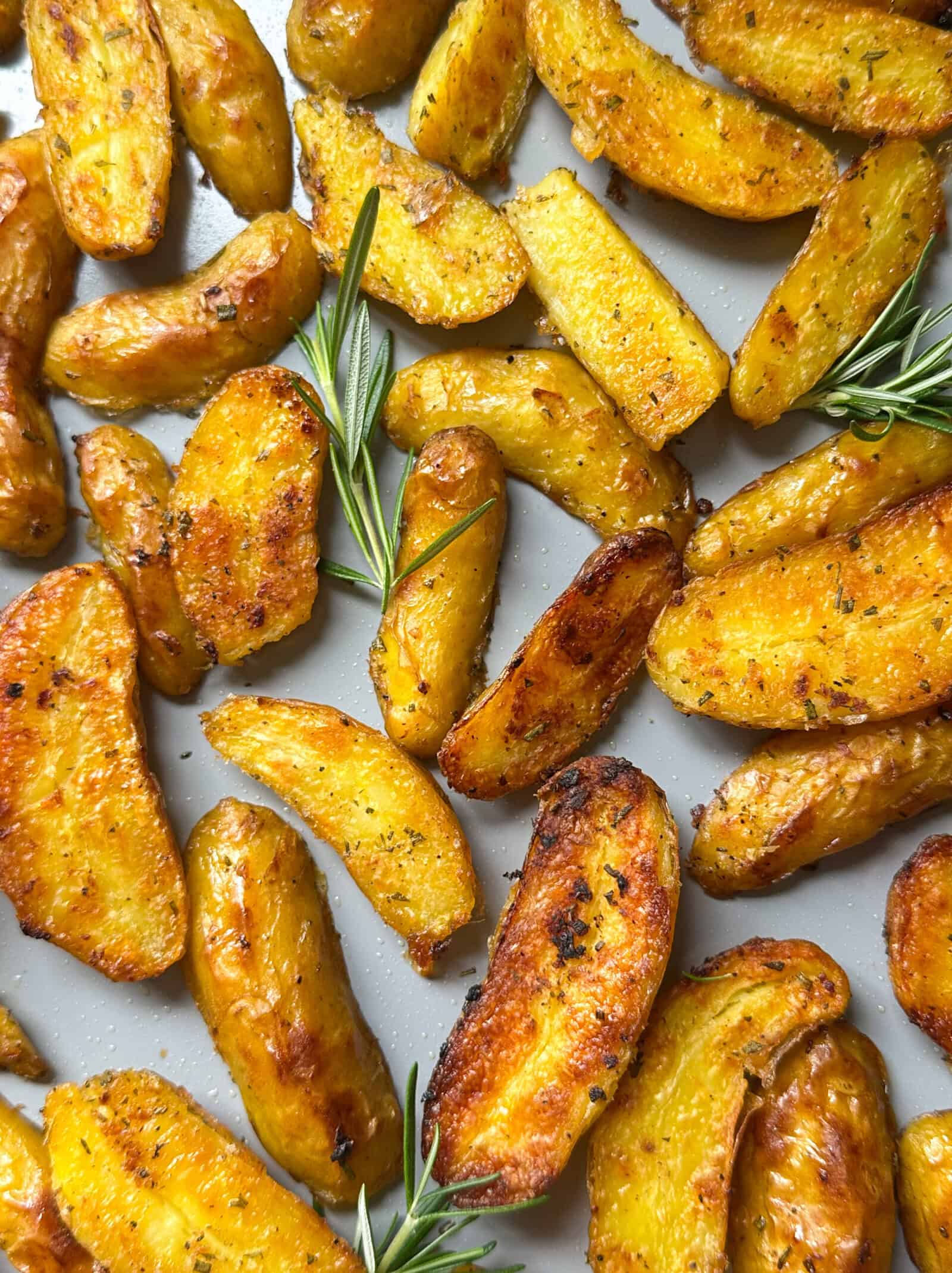Golden, crispy roasted potatoes are scattered on a baking tray, garnished with fresh rosemary. Perfectly seasoned with a slightly charred look, they’re an irresistible example of how to make crispy roasted potatoes at home.