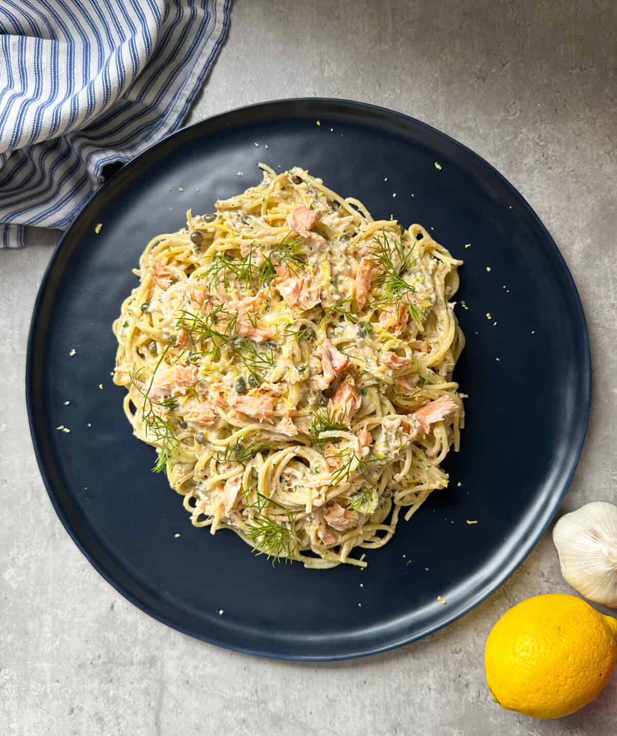 A dark plate holds creamy salmon pasta, topped with fresh dill and succulent salmon. Nearby are a striped cloth, a lemon, and a head of garlic on a light gray surface—an inviting scene for any salmon pasta recipe lover.