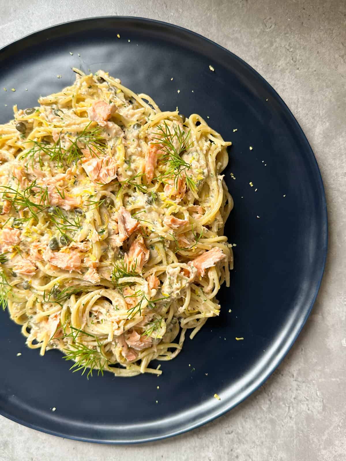 Spaghetti topped with creamy sauce, flaked salmon, fresh dill, and lemon zest—this creamy salmon pasta is served on a dark blue plate on a light-colored surface.