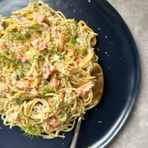 A dark plate with creamy salmon pasta topped with chunks of salmon and fresh dill, with a spoon resting on the plate.