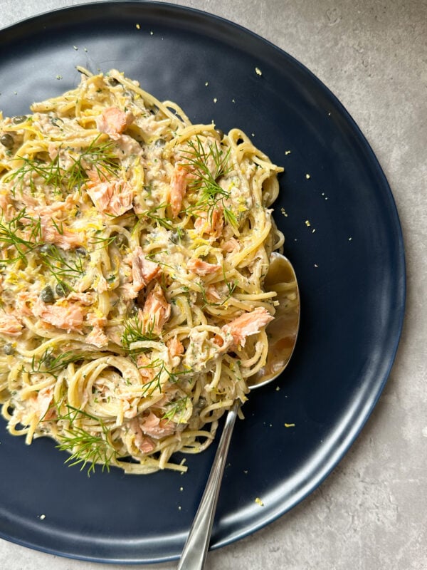 A dark plate with creamy salmon pasta topped with chunks of salmon and fresh dill, with a spoon resting on the plate.