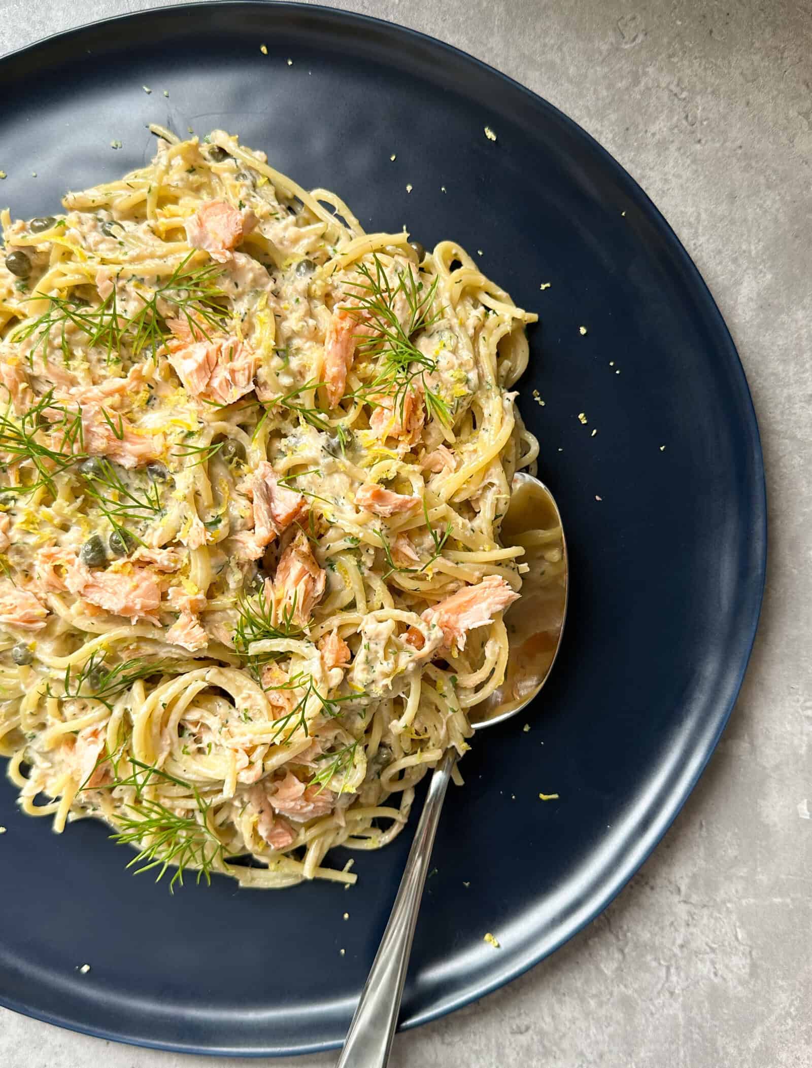A dark plate with creamy salmon pasta topped with chunks of salmon and fresh dill, with a spoon resting on the plate.