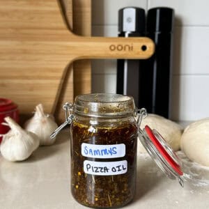 A glass jar labeled SAMMYS PIZZA OIL sits on a kitchen counter, surrounded by garlic bulbs, pizza dough, oil bottles, and a wooden pizza board—perfect for drizzling over hot oil pizza or trying your favorite pizza oil recipe.
