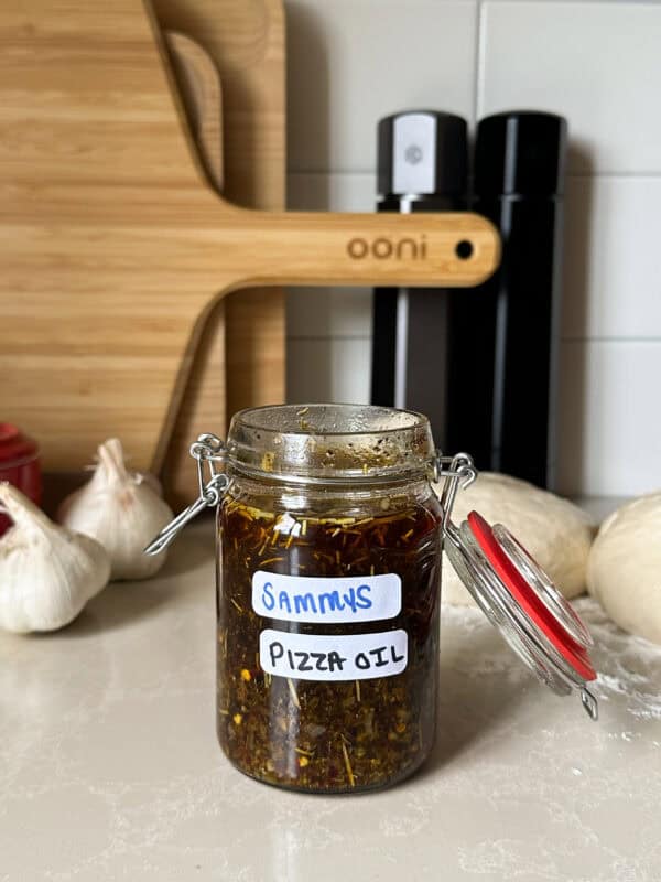A glass jar labeled SAMMYS PIZZA OIL sits on a kitchen counter, surrounded by garlic bulbs, pizza dough, oil bottles, and a wooden pizza board—perfect for drizzling over hot oil pizza or trying your favorite pizza oil recipe.