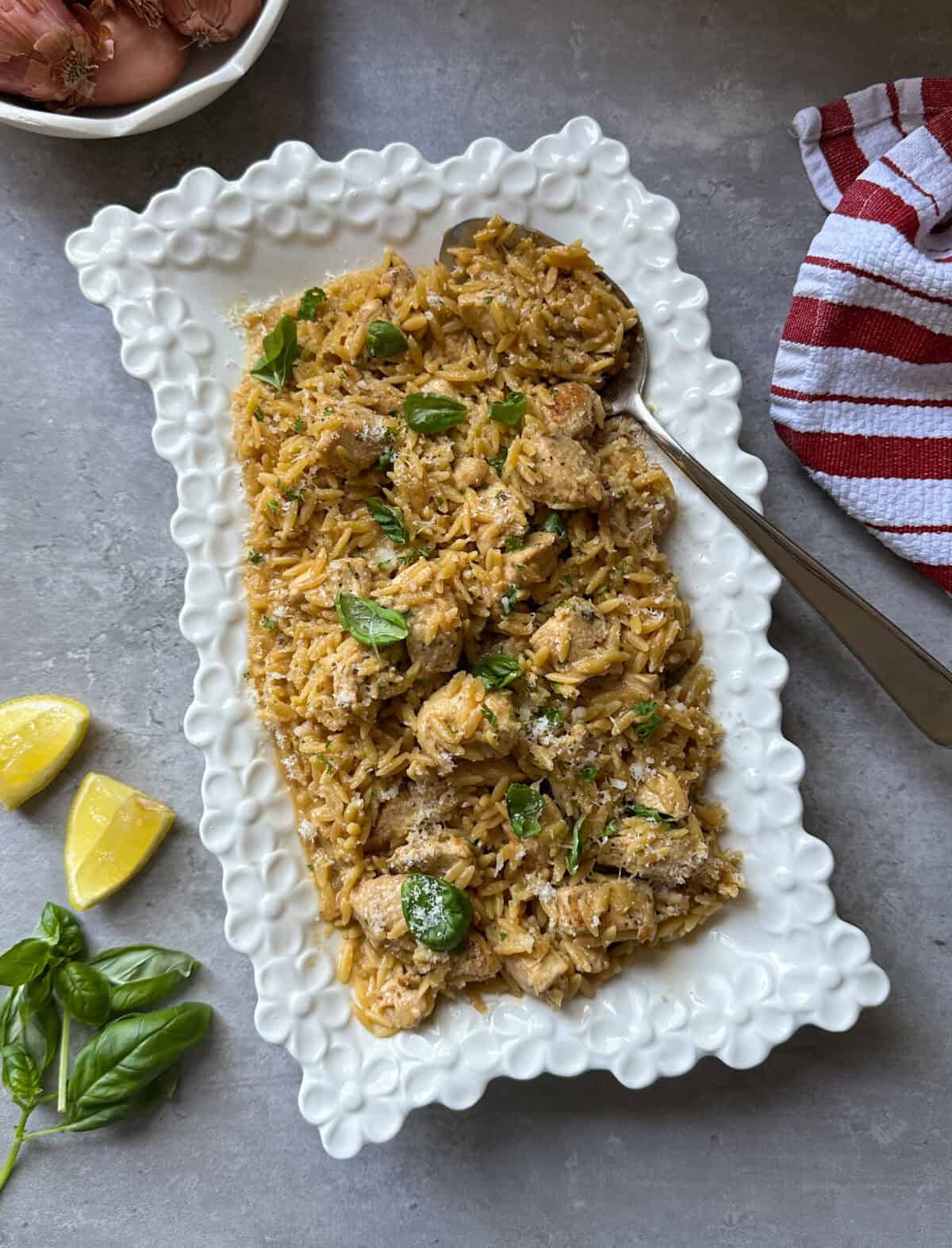 A white decorative platter filled with lemon chicken orzo is garnished with fresh basil. A spoon rests on the platter, while lemon wedges, basil leaves, shallots, and a red-striped towel are arranged nearby on a gray surface.