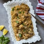 A white decorative platter filled with lemon chicken orzo is garnished with fresh basil. A spoon rests on the platter, while lemon wedges, basil leaves, shallots, and a red-striped towel are arranged nearby on a gray surface.