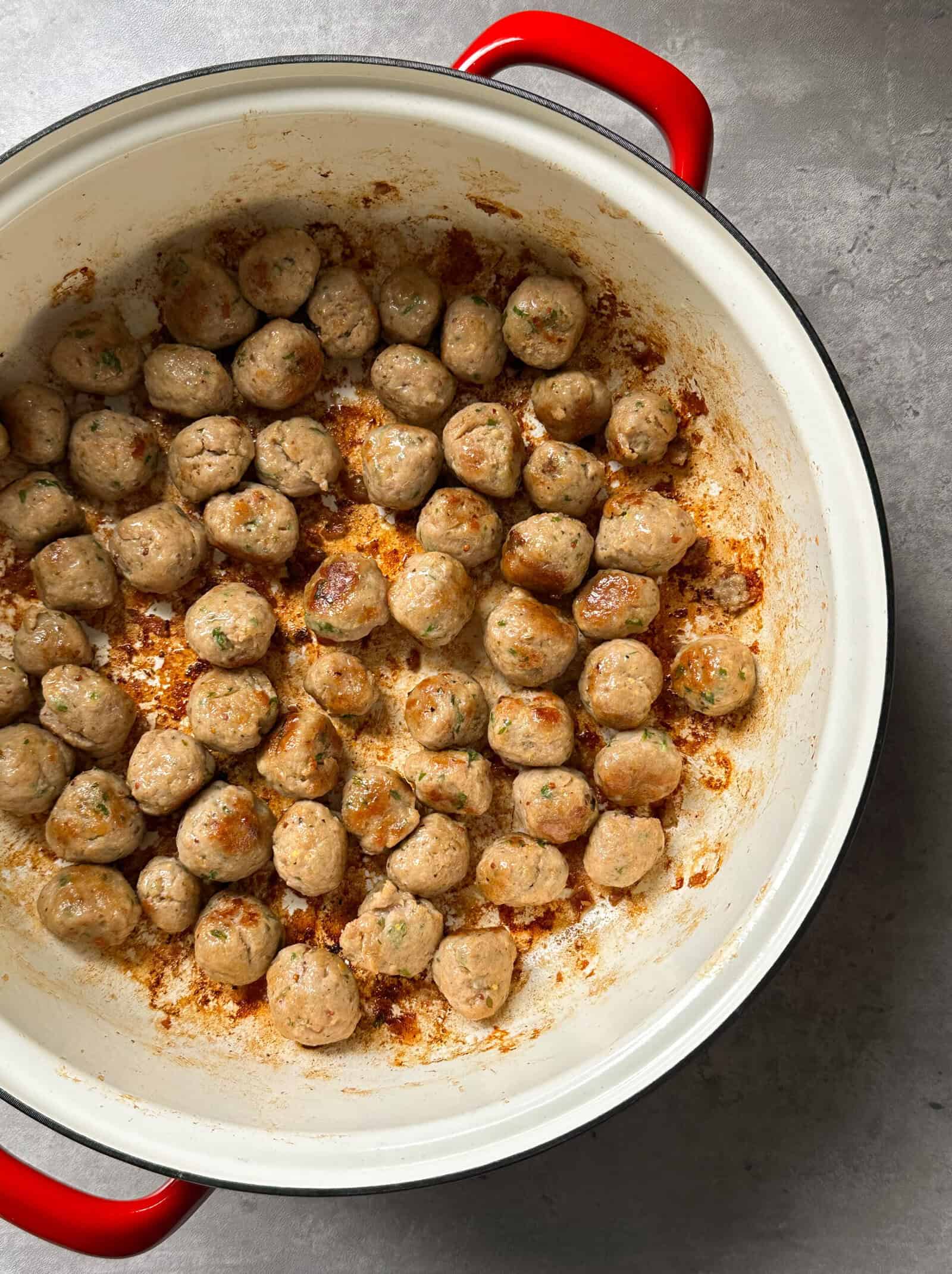 A white pot with red handles holds browned meatballs scattered across the bottom, with tasty browned bits—an inviting base for a creamy Tuscan chicken pasta recipe. The background features a simple gray surface.