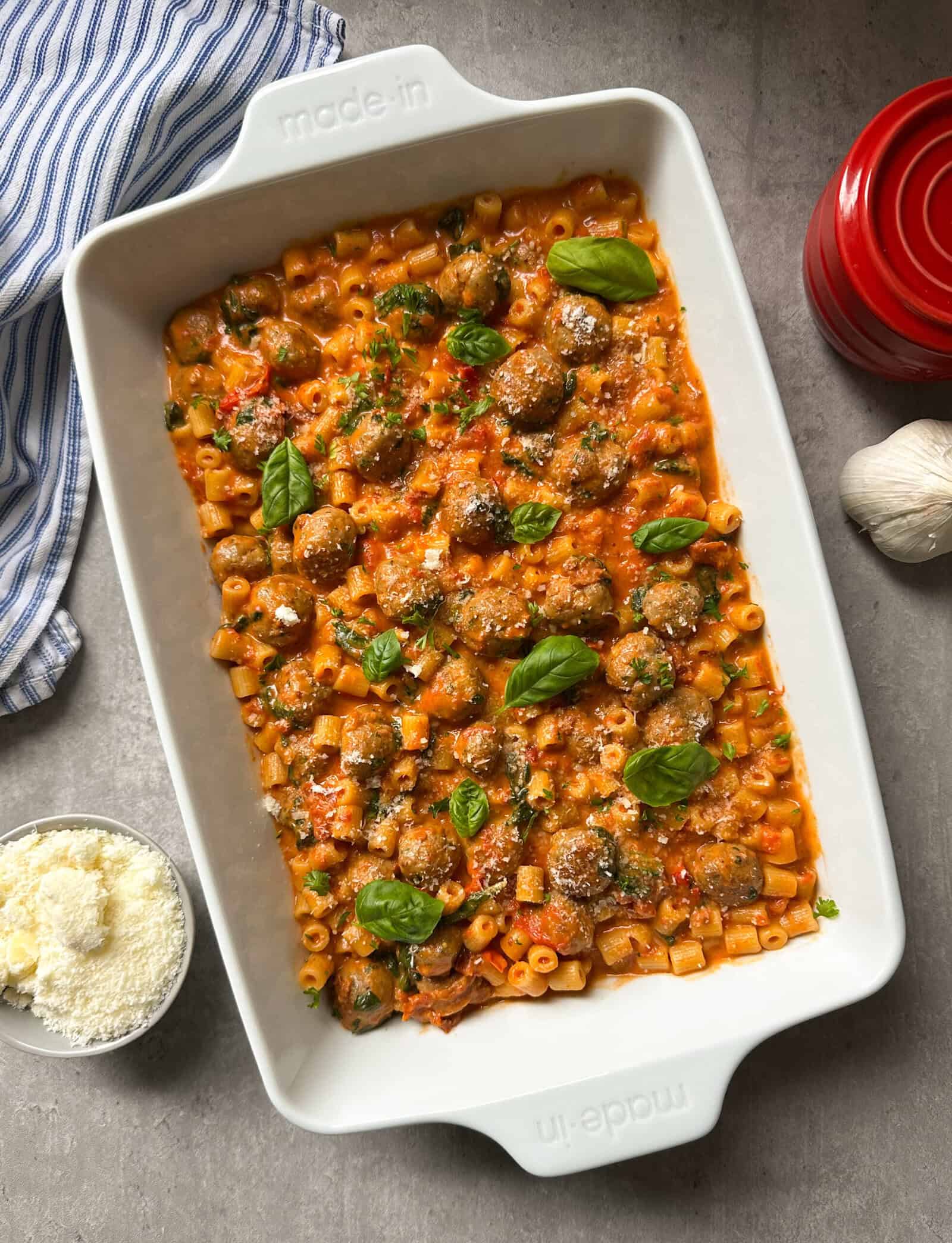 A white baking dish filled with tuscan chicken pasta and meatballs in a creamy tomato sauce, garnished with fresh basil. Nearby are a blue-striped towel, a red container, garlic, and a bowl of grated cheese.